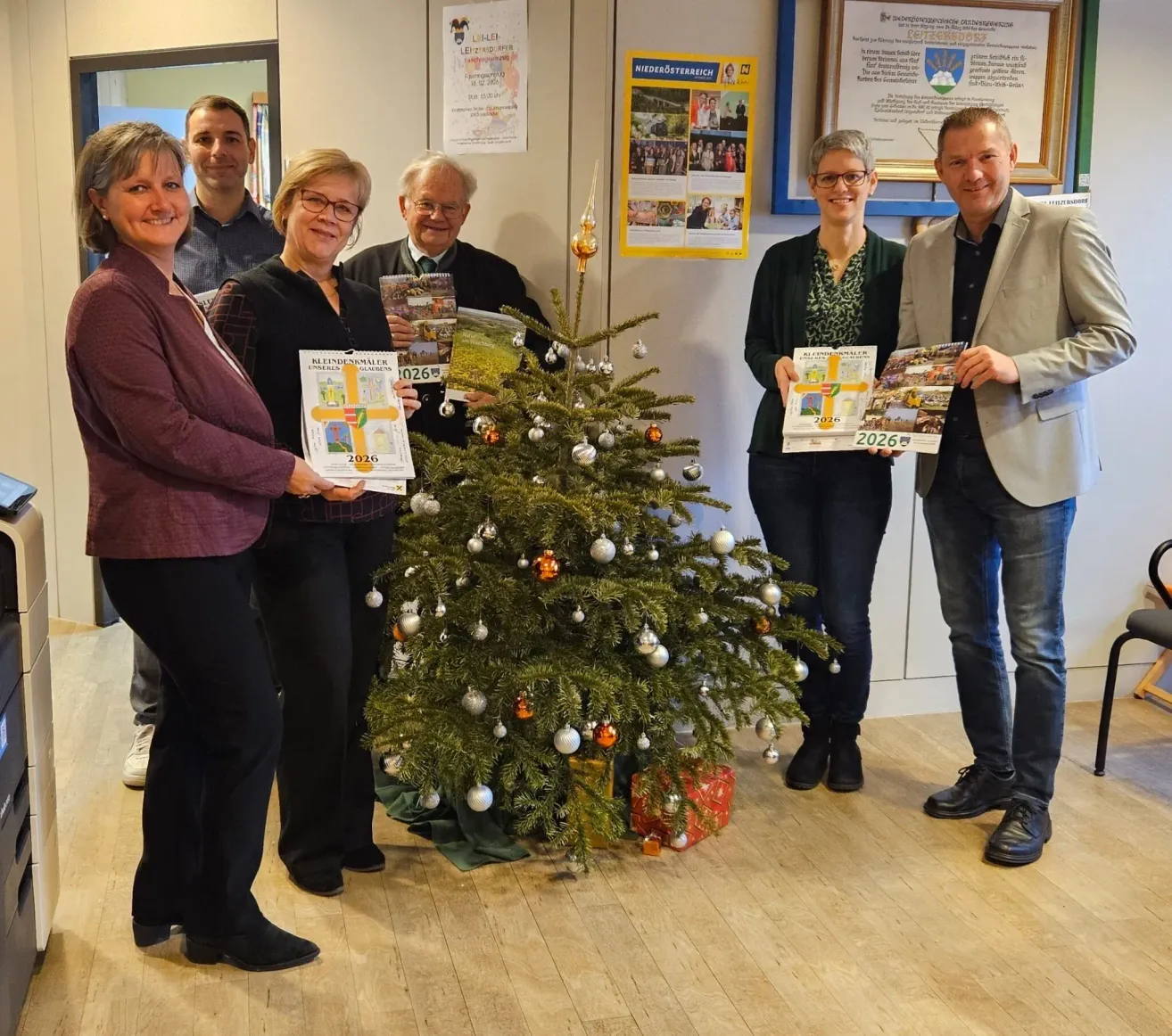 Fünf Personen stehen um einen geschmückten Weihnachtsbaum herum und halten Bücher mit 2026 auf dem Cover. Dahinter befindet sich eine Wand mit Postern und einem großen Rahmen.