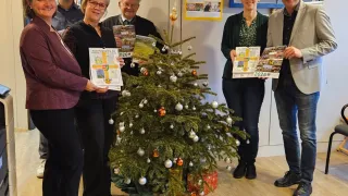 Fünf Personen stehen um einen geschmückten Weihnachtsbaum herum und halten Bücher mit 2026 auf dem Cover. Dahinter befindet sich eine Wand mit Postern und einem großen Rahmen.