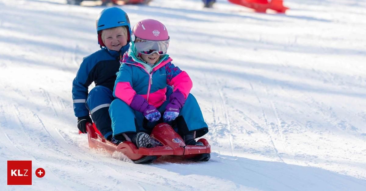 Zwei Kinder sitzen auf einem roten Schlitten, tragen Winterkleidung und Helme. Sie lächeln und genießen einen verschneiten Tag.