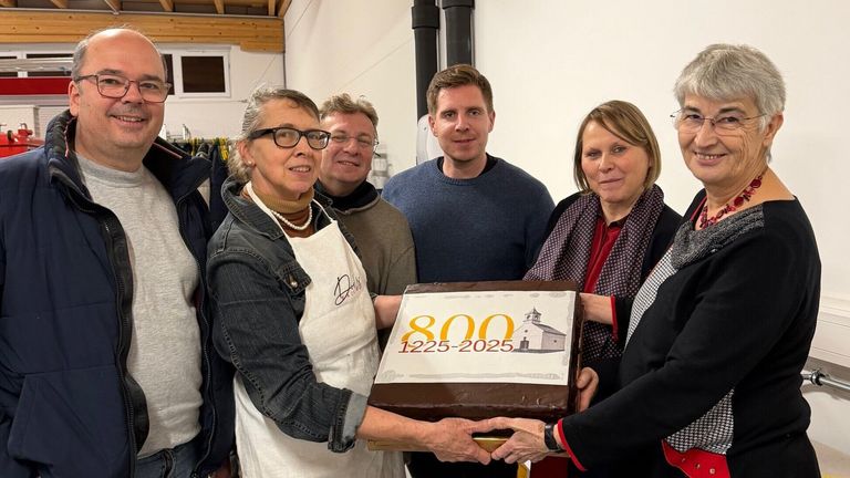 Fünf Menschen stehen um einen Kuchen mit '800 1225-2025' herum. Die Frau in der weißen Schürze hält den Kuchen. Der Mann im Pullover lächelt, und die anderen schauen zu. Sie scheinen einen Meilenstein zu feiern.