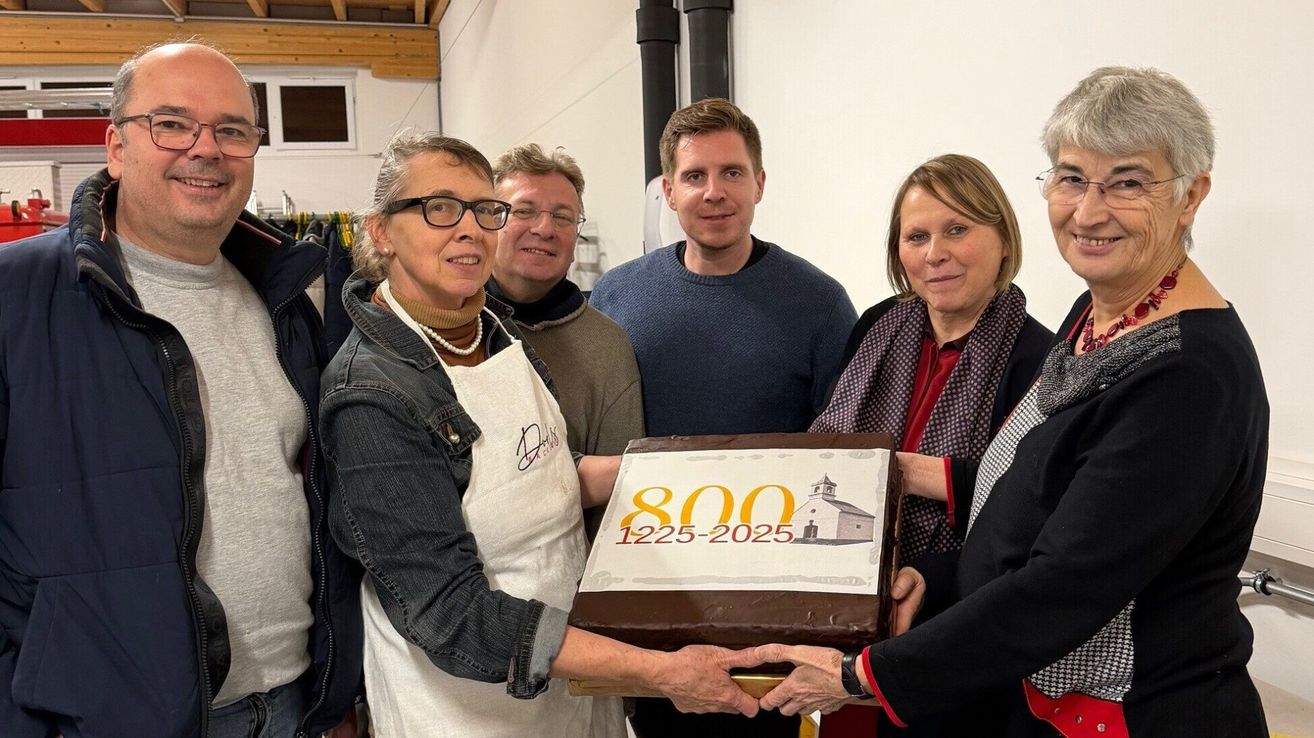 Fünf Menschen stehen um einen Kuchen mit '800 1225-2025' herum. Die Frau in der weißen Schürze hält den Kuchen. Der Mann im Pullover lächelt, und die anderen schauen zu. Sie scheinen einen Meilenstein zu feiern.