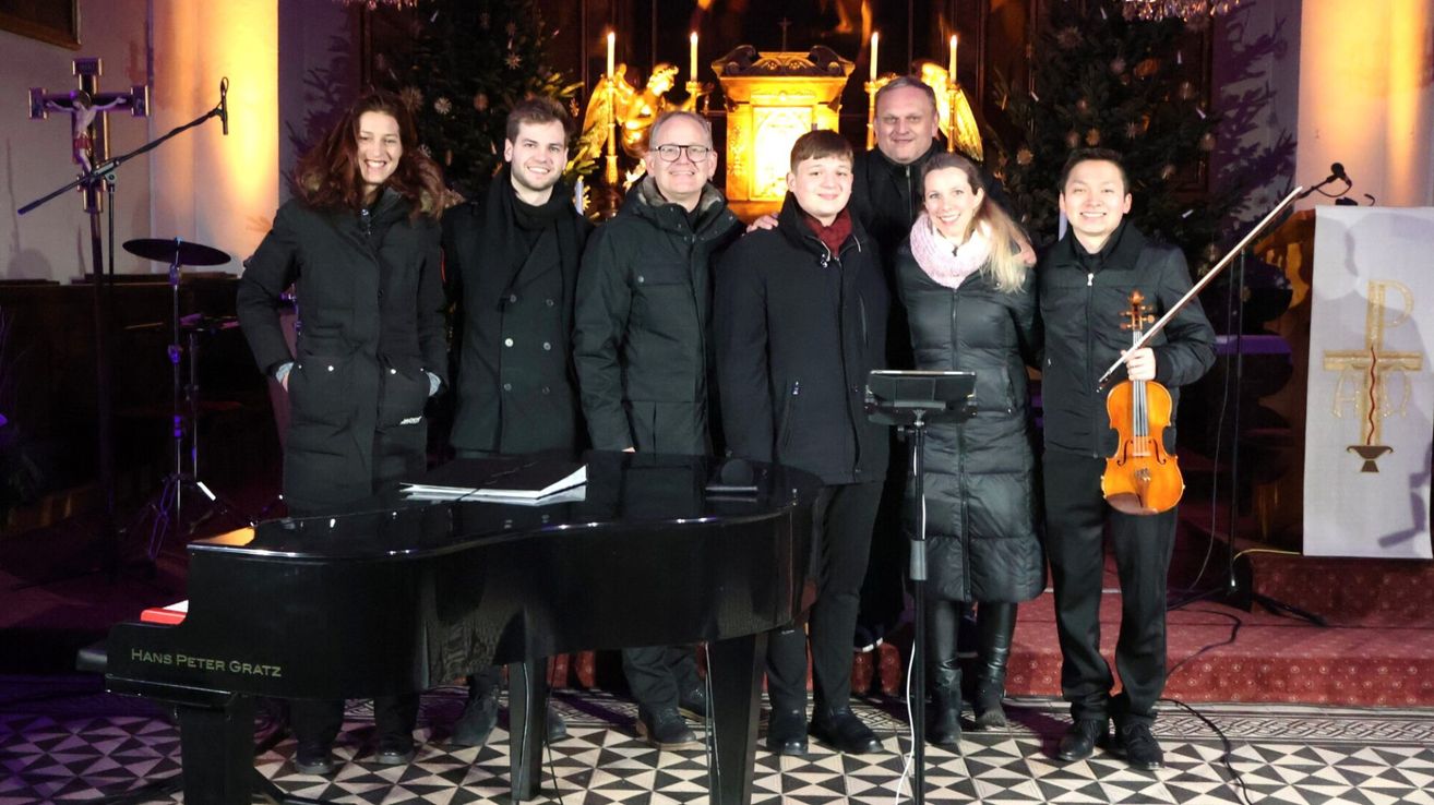 Eine Gruppe von Menschen in schwarzen Mänteln steht hinter einem Klavier und einem Notenständer. Sie befinden sich vor einem dekorierten Kirchenbereich mit Kerzen und einer goldenen Struktur. Sie scheinen sich auf eine Aufführung vorzubereiten.