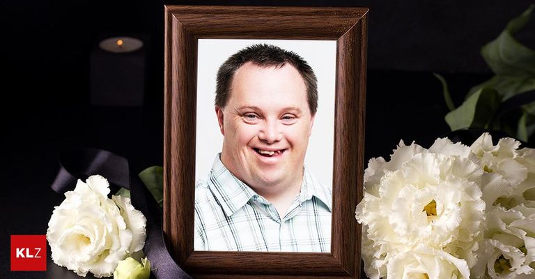 Ein hölzerner Rahmen mit einem lächelnden Mann in einem karierten Hemd, umgeben von weißen Blumen und einer Kerze.