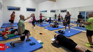 Eine Gruppe von Menschen in einem Fitnessstudio, einige üben mit Hanteln und andere mit einer Stange. Sie tragen Sportbekleidung und es liegen Matten und Übungsbälle auf dem Boden.