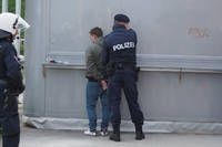 Zwei Polizeibeamte stehen neben einer Wand, einer von ihnen nimmt einen Mann in einem grünen Jackett fest. Der Mann hat seine Hände auf dem Rücken.