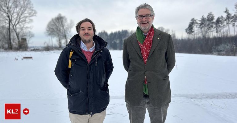Zwei Männer stehen in einem verschneiten Feld und lächeln. Der Mann auf der linken Seite trägt eine schwarze Jacke und einen gelben Riemen. Der Mann auf der rechten Seite trägt einen grauen Anzug und einen rot-grün gemusterten Schal. Bäume sind im Hintergrund zu sehen.