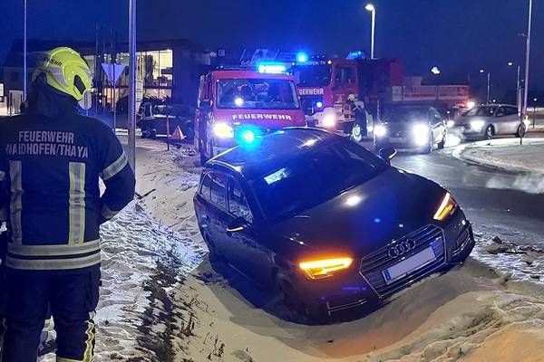 Feuerwehrleute und Rettungsfahrzeuge kümmern sich um einen schwarzen Audi auf einer verschneiten Straße, wobei das Auto zur Seite geneigt ist. Ein Feuerwehrmann steht in der Nähe des Autos, und mehrere andere Autos bewegen sich auf der Straße.