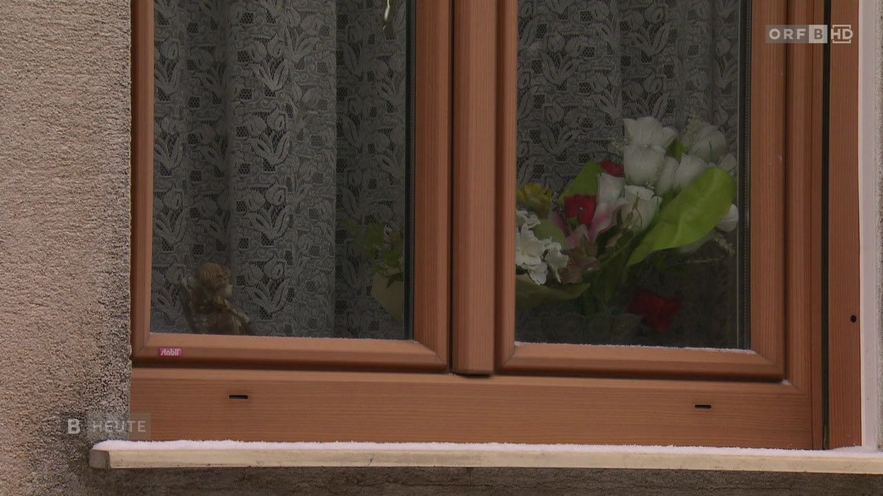 Ein hölzernes Fenstergestell mit Blumenvorhängen, ein Teddybär auf der linken Seite sichtbar und ein Blumenstrauß auf der rechten Seite.