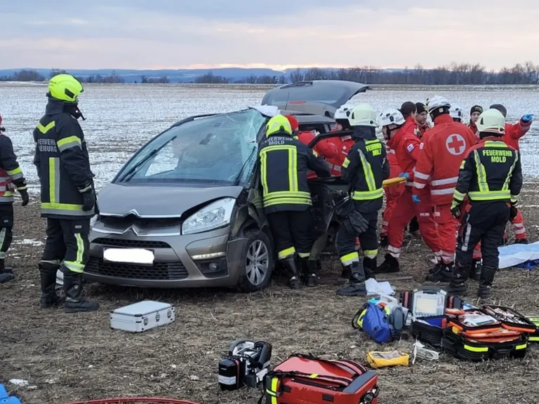 Rettungskräfte in Schutzausrüstung kümmern sich um ein beschädigtes Fahrzeug. Das Auto hat eine eingeschlagene Frontscheibe. Medizinisches Equipment und Werkzeuge sind verstreut.