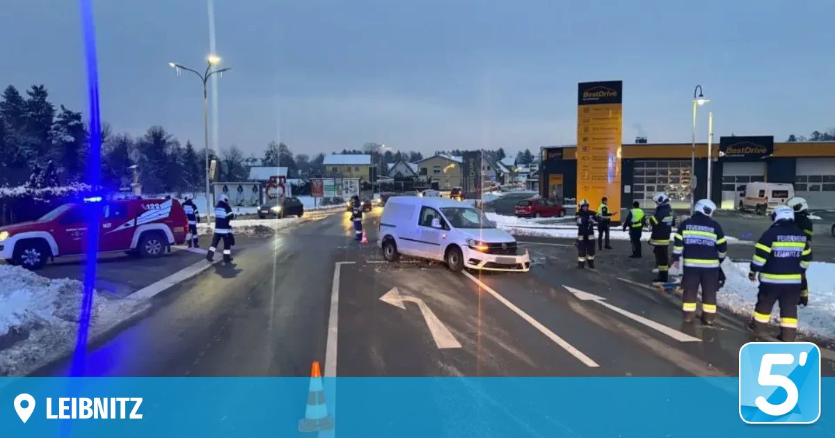 Rettungskräfte leiten den Verkehr in der Nähe eines Unfalls mit einem weißen Lieferwagen auf einer verschneiten Straße mit Gebäuden und einem Schild im Hintergrund.