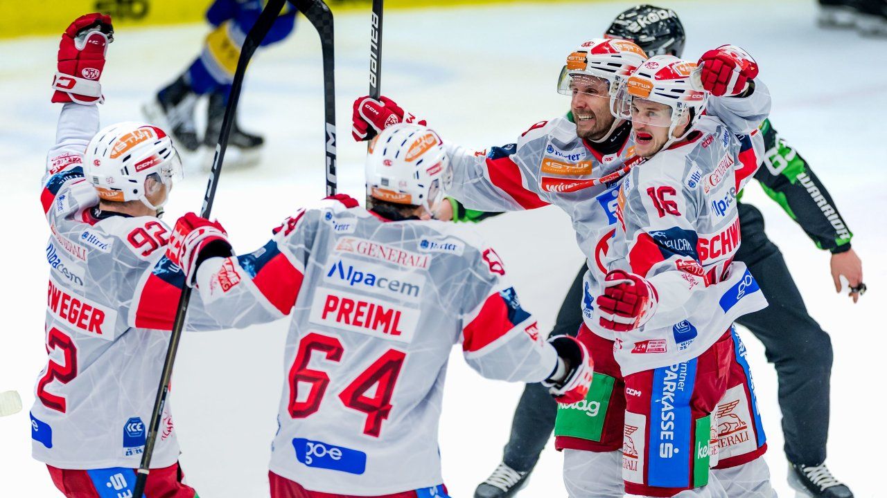 Eishockeyspieler in weißen und roten Trikots feiern ein Tor auf der Eisbahn. Die Spieler halten Eishockeyschläger und tragen Helme. Die Trikots zeigen verschiedene Sponsorenlogos.