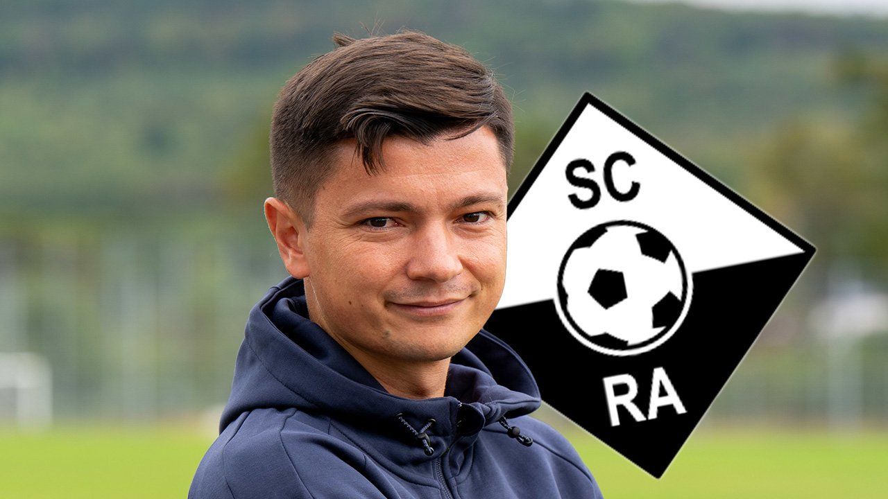 Ein Mann mit einer blauen Kapuzenjacke lächelt vor einem SC RA Fußballlogo.
