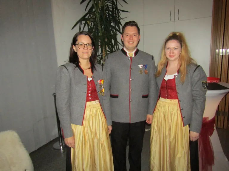 Drei Menschen in traditioneller Kleidung mit Medaillen um den Hals posieren für ein Foto. Dahinter befindet sich eine weiße Wand und eine Pflanze.