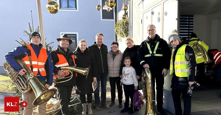 Eine Gruppe von Erwachsenen und Kindern steht draußen und lächelt für ein Foto. Zwei Männer halten Messinginstrumente und ein Mädchen hat einen Rucksack. Das Gebäude hat Fenster und Weihnachtsdekoration.
