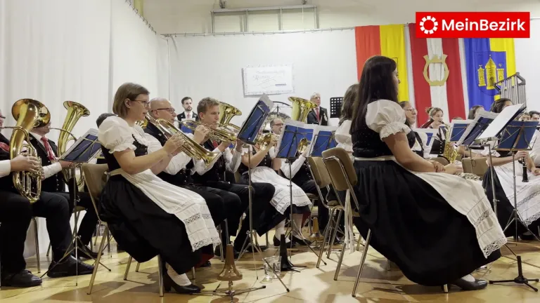 Eine Bläsergruppe spielt im Innenbereich. Die Bandmitglieder tragen traditionelle Kleidung. Das Publikum sitzt auf Stühlen. Eine Flagge hängt an der Wand.