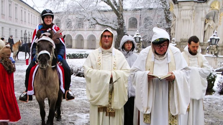 Eine verschneite Szene mit Priestern in weißen Roben. Einer liest ein Buch, und ein anderer hält einen Stock. Ein Mensch auf einem Pferd in einer Weihnachtsmann-Kostümierung ist in der Nähe. Dahinter befindet sich ein Gebäude mit Bogenfenstern.