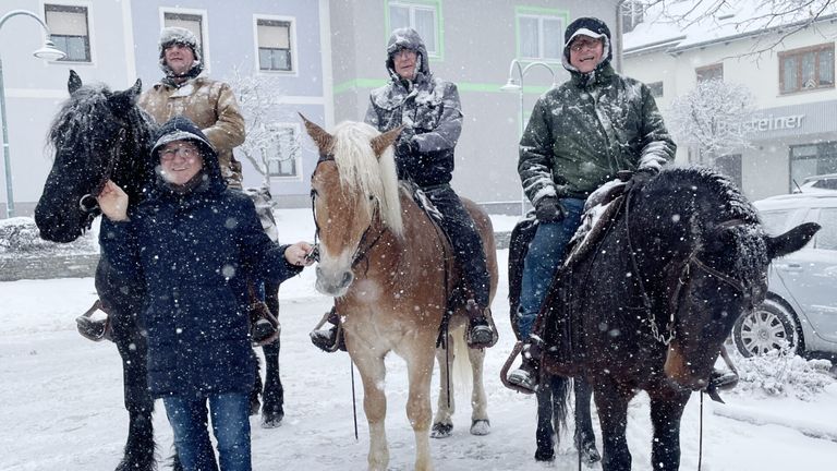 Vier Personen in Winterkleidung stehen im Schnee mit ihren Pferden. Drei reiten, während die vierte die Zügel eines Pferdes hält. Die Pferde sind schneebedeckt, und ein Gebäude mit grüner Umrandung ist im Hintergrund zu sehen.