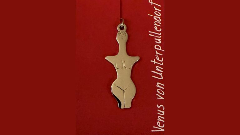 Ein goldener Anhänger in Form einer Venusfigur hängt an einer Kette vor rotem Hintergrund. Der Text lautet 'Venus von Unterpullendorf'.