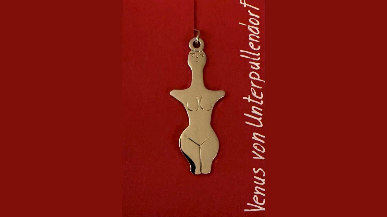 Ein goldener Anhänger in Form einer Venusfigur hängt an einer Kette vor rotem Hintergrund. Der Text lautet 'Venus von Unterpullendorf'.