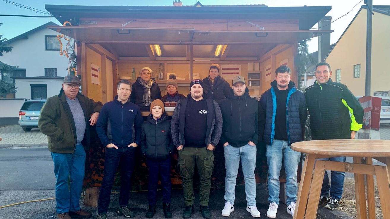Eine Gruppe von Menschen in Winterkleidung steht vor einer Holzstruktur, möglicherweise einem Food Truck, mit Regalen und Gegenständen im Inneren. Sie scheinen für ein Foto zu posieren.