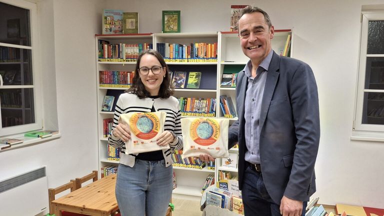 Eine Frau und ein Mann stehen in einer Bibliothek mit Regalen voller Bücher. Die Frau hält eine Tasche mit dem Wort Burgenland, und der Mann hält eine ähnliche Tasche.