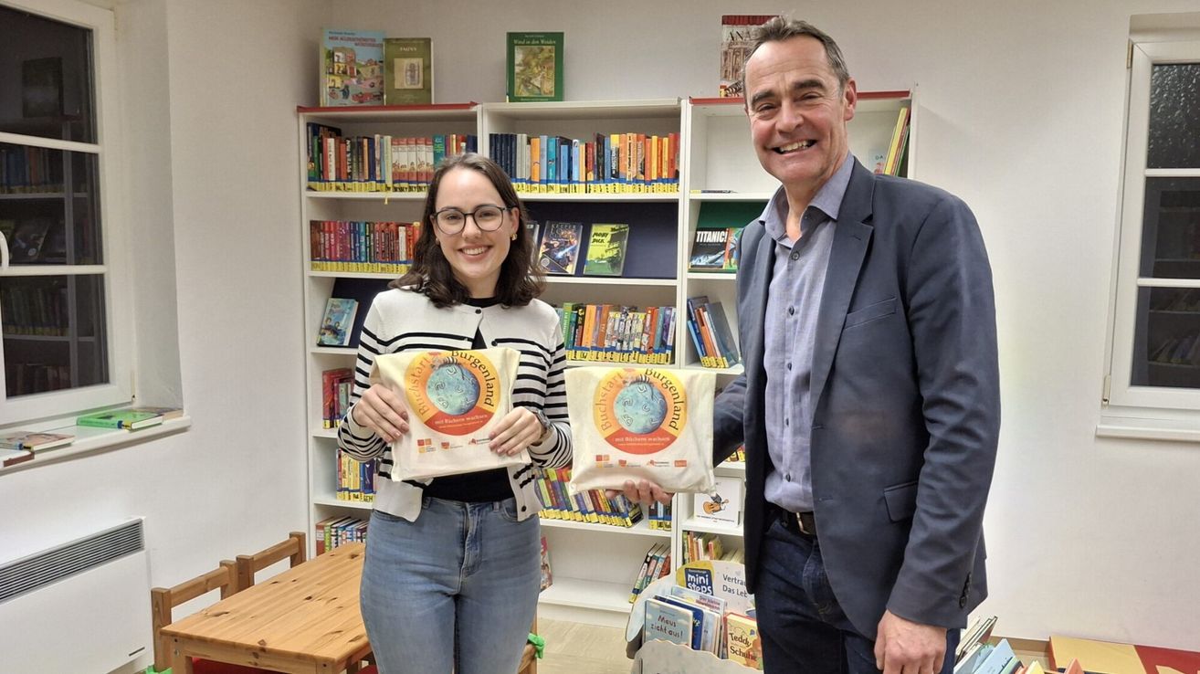 Eine Frau und ein Mann stehen in einer Bibliothek mit Regalen voller Bücher. Die Frau hält eine Tasche mit dem Wort Burgenland, und der Mann hält eine ähnliche Tasche.