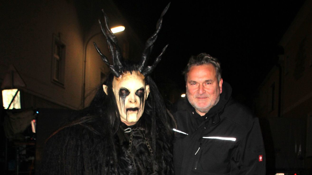 Ein Mann in einer schwarzen Jacke steht neben einer Person in einer gruseligen hörnigen Maske und schwarzem Kostüm, möglicherweise bei Nacht.