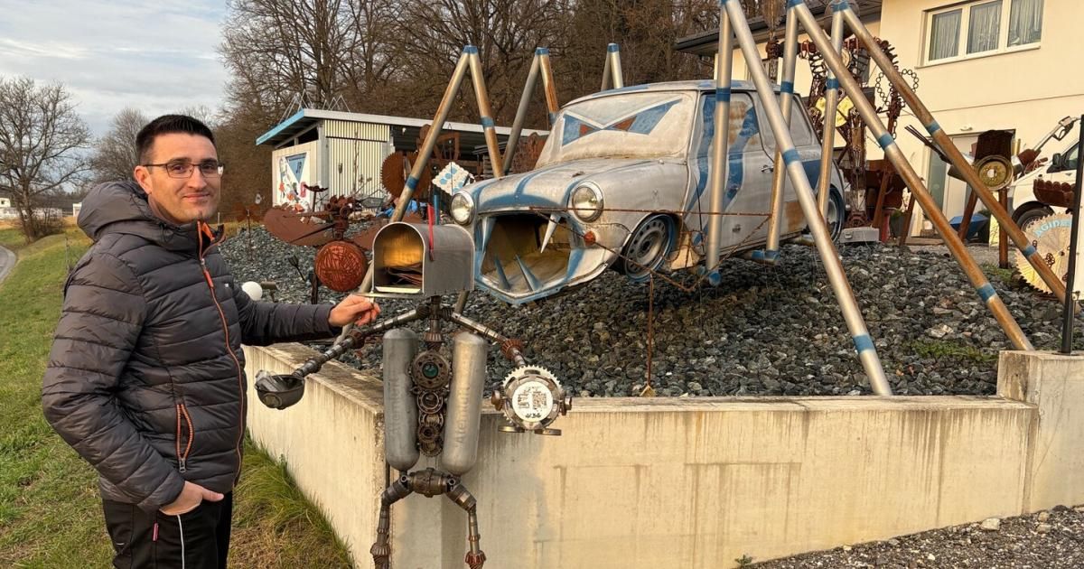 Ein Mann posiert mit einer mechanischen Roboterskulptur, einem angebauten Briefkasten und einem zerbrochenen Auto, das auf Kies neben einem Gebäude thront.
