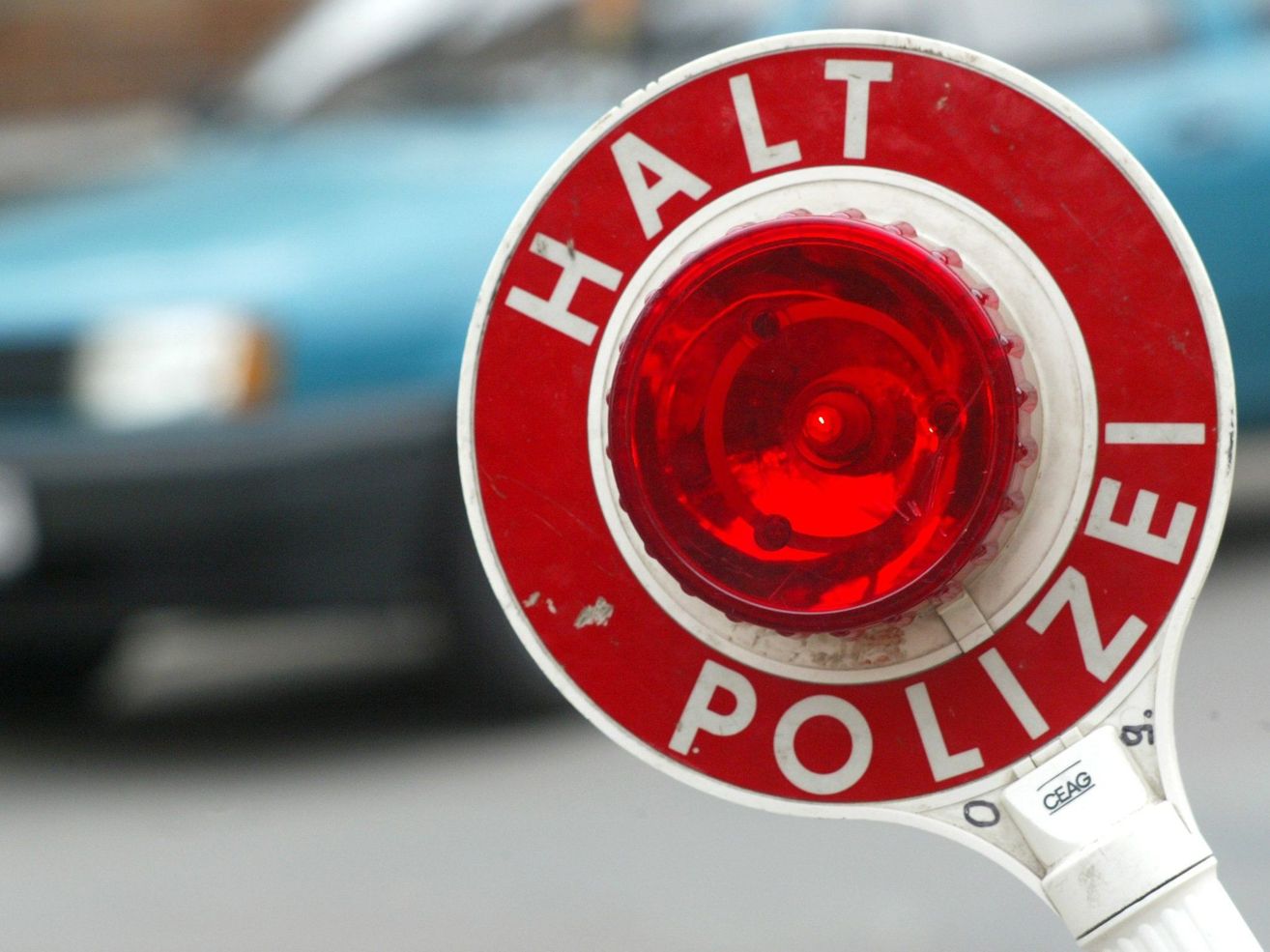 Nahaufnahme einer roten blinkenden Polizei-Stopp-Leuchte mit dem Wort 'HALT' in Weiß und 'POLIZEI' in Rot. Die Leuchte ist an einem weißen Griff befestigt. Im Hintergrund befindet sich ein blaues Auto.