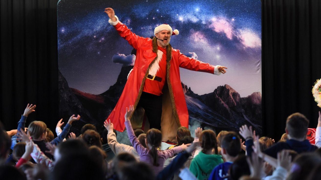 Ein Mann in einer Santa-Claus-Verkleidung tritt auf der Bühne auf, vor einem Hintergrund mit Sternenhimmel. Kinder schauen ihm begeistert zu.