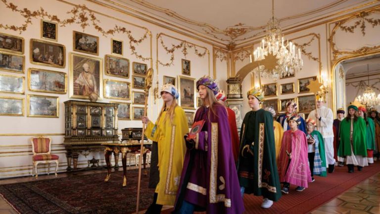 Eine Gruppe von Kindern in traditionellen Kostümen geht durch einen prunkvollen Raum, geschmückt mit Gemälden und einer Kristallkronleuchter. Die Kinder tragen farbenfrohe Hüte und Umhänge.