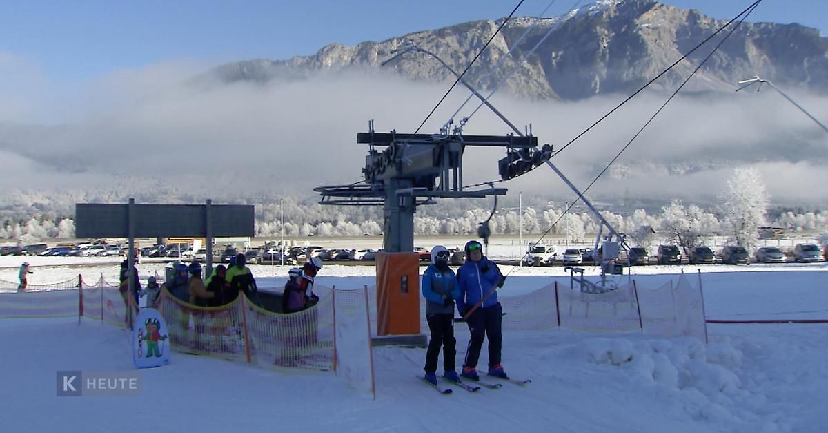 Zwei Skifahrer posieren für ein Foto an einem Skilift mit einem Berg im Hintergrund.