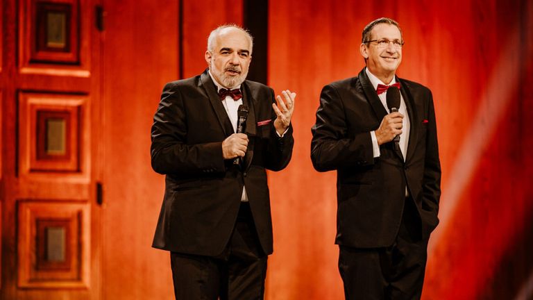 Zwei Männer in Anzügen stehen auf der Bühne, einer hält ein Mikrofon und gestikuliert, der andere lächelt und hält ein Mikrofon mit einer roten Fliege.