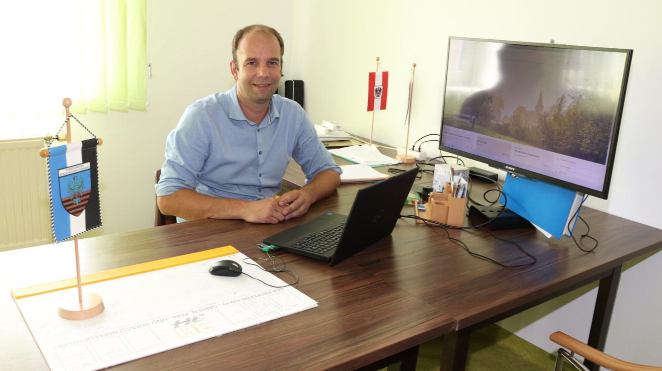 Ein Mann sitzt an einem Schreibtisch mit einem Laptop, einer Flagge dahinter und einem Monitor auf dem Schreibtisch. Papiere, eine Maus und ein Stift liegen auf dem Schreibtisch.
