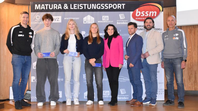 Fünf Personen stehen auf einer Bühne, halten Urkunden in der Hand, vor einem Hintergrund mit Logos und den Worten 'Natura & Leistungssport.' Sie scheinen bei einer Preisverleihung zu sein.
