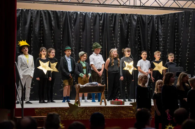 Eine Gruppe von Kindern in Kostümen steht auf der Bühne und führt ein Weihnachtsspiel auf. Sie singen mit einem Mikrofon vor sich. Die Bühne hat einen Hintergrund aus einem schwarzen Vorhang.