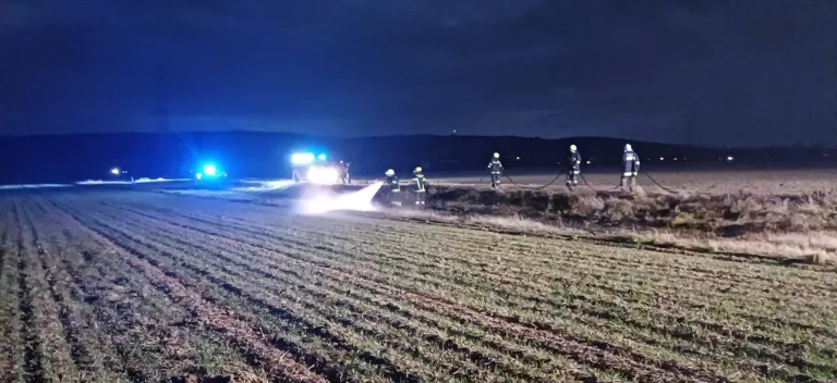 Firefighters in full gear are battling a blaze at night in a large field. A fire truck is parked nearby, and a hose is being used to spray water.