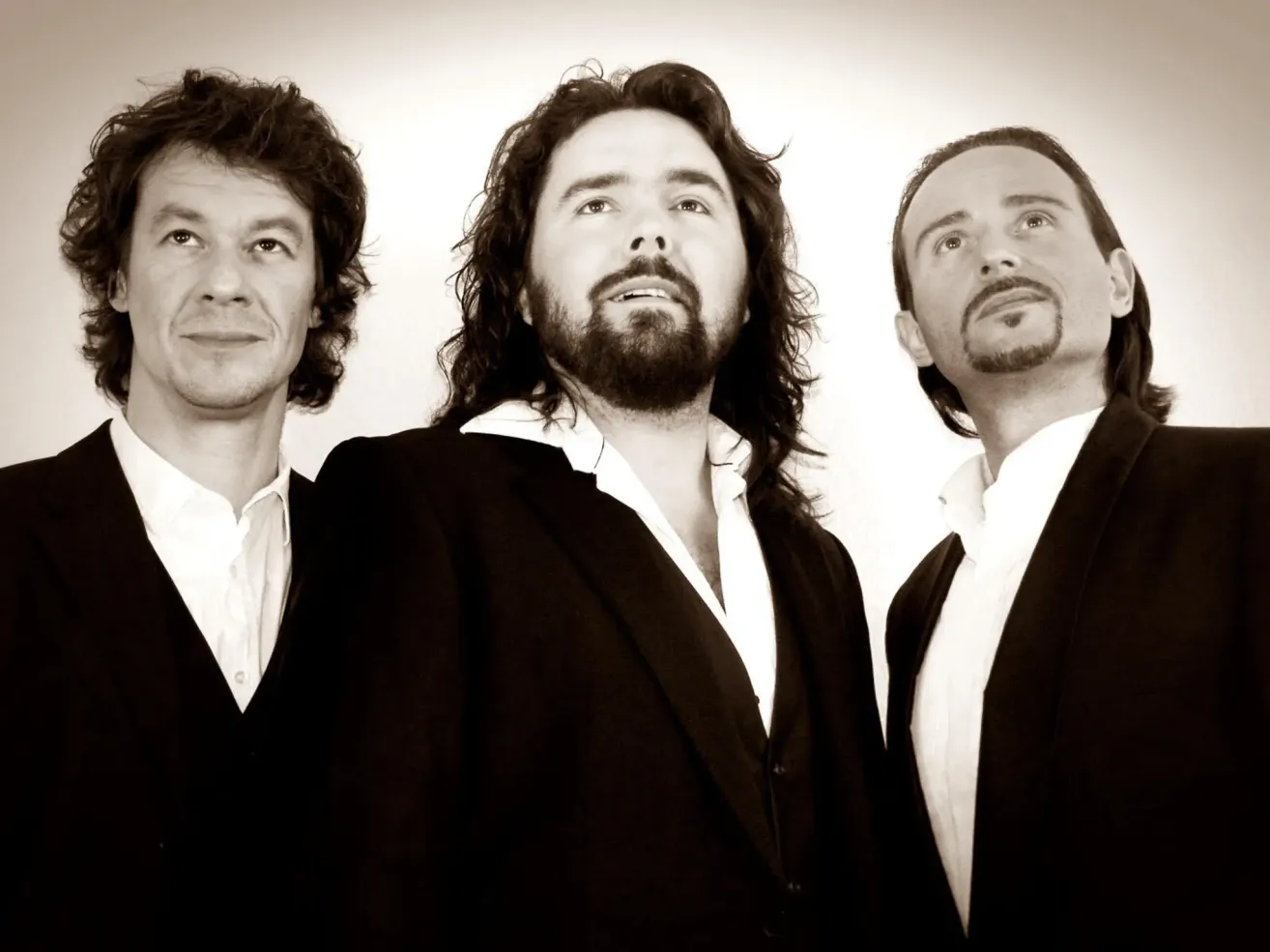 Drei Männer in schwarzen Anzügen mit weißen Hemden posieren für ein Schwarz-Weiß-Foto. Der Mann in der Mitte hat lange Haare und einen Bart.