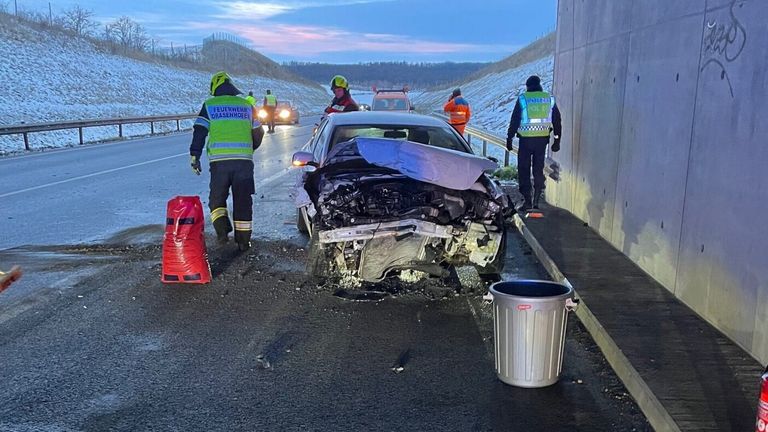 Ein schwer beschädigtes Auto steht am Straßenrand mit anwesendem Notfallpersonal. Zwei Personen in reflektierenden Westen und Helmen sind in der Nähe.