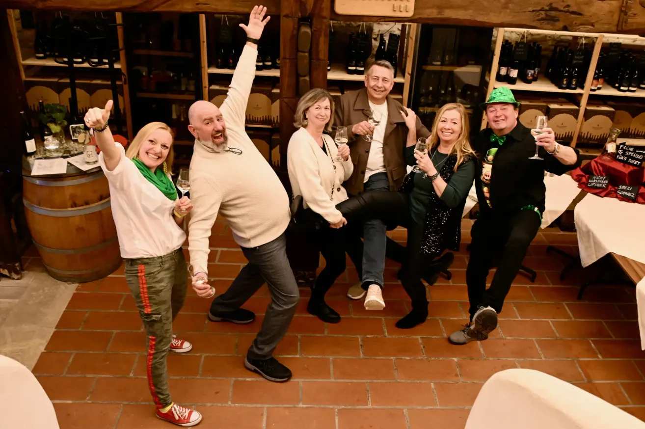 Sechs Menschen posieren für ein Foto in einem Keller und halten Weingläser in den Händen. Sie alle lächeln und scheinen zu feiern. Der Keller ist mit Regalen voller Weinflaschen gefüllt.