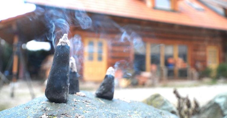 Ein Nahaufnahme von drei brennenden Räucherstäbchen auf einem Felsen, die Rauch abgeben vor einem Holzhaus. Das Haus hat eine gelbe Tür und Fenster.