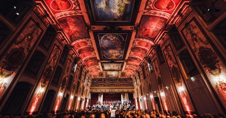 Ein großer Saal mit einem Orchester, das auf der Bühne auftritt. Die Decke und die Wände sind mit kunstvollen Bildern und roter Beleuchtung geschmückt. Das Publikum sitzt vor der Bühne.