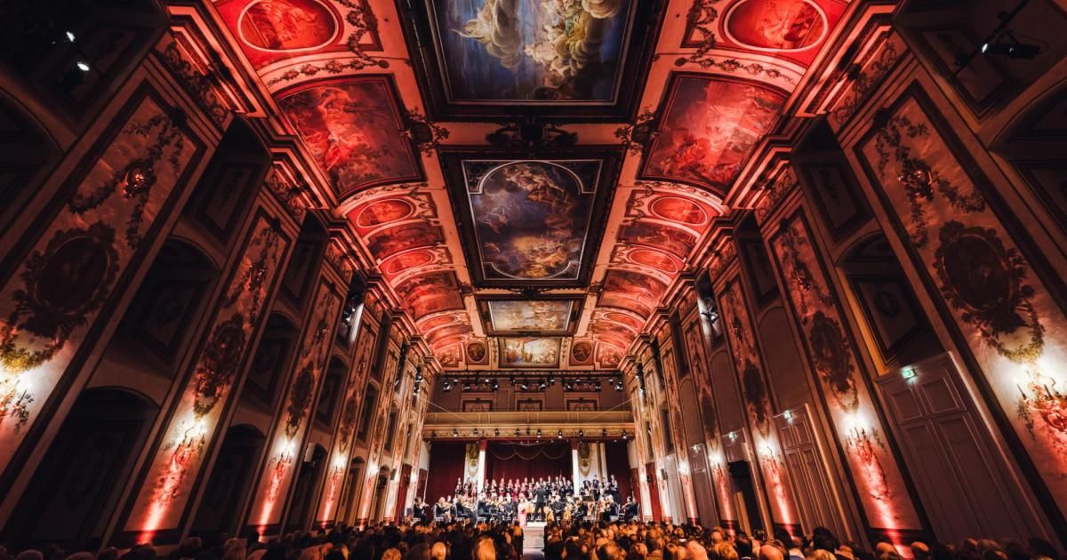 Ein großer Saal mit einem Orchester, das auf der Bühne auftritt. Die Decke und die Wände sind mit kunstvollen Bildern und roter Beleuchtung geschmückt. Das Publikum sitzt vor der Bühne.