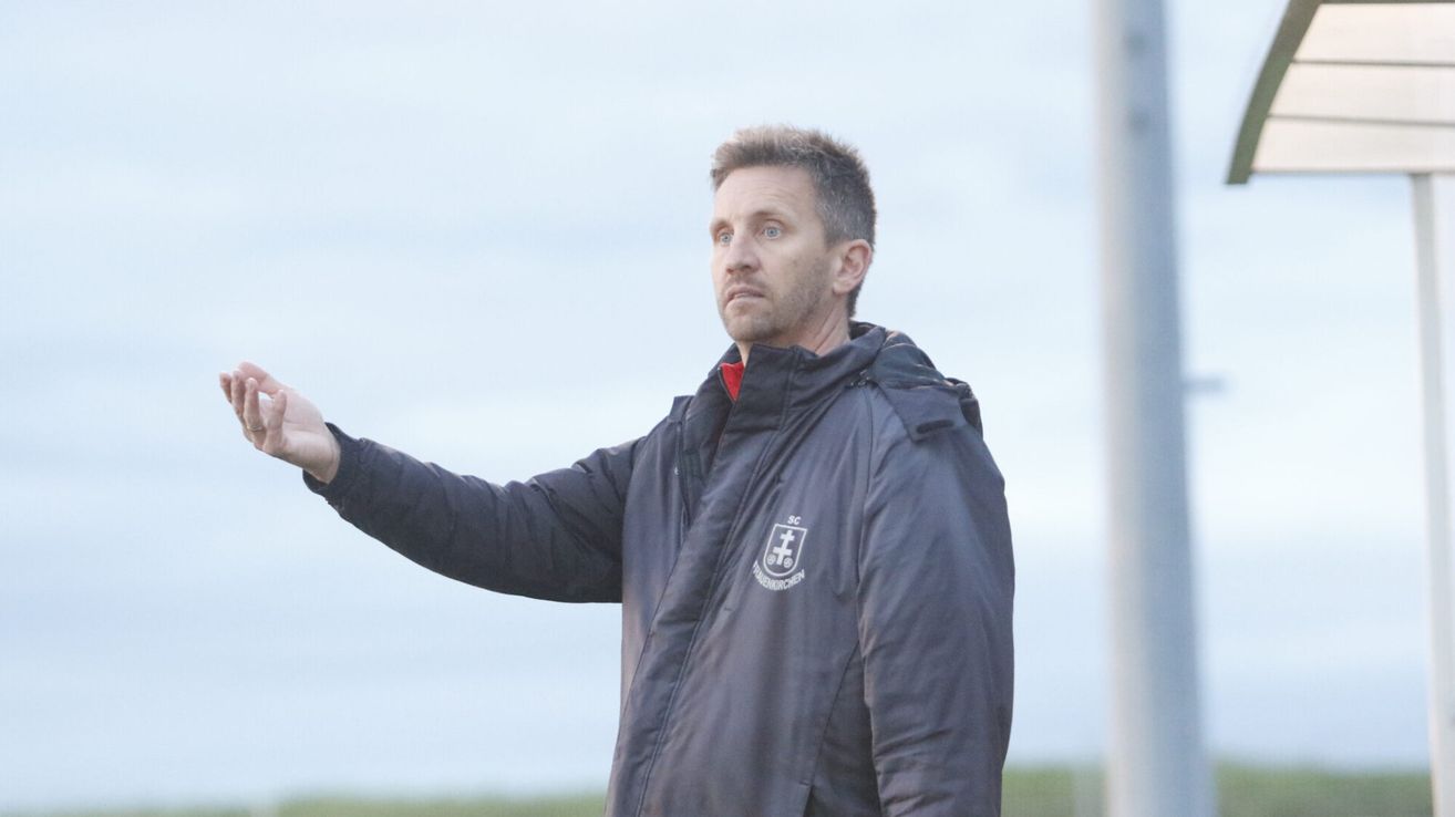 Ein Mann steht im Freien und trägt eine dunkle Jacke mit einem roten T-Shirt darunter. Er zeigt nach links und sieht ernst aus. Im Hintergrund deutet ein verschwommener Himmel auf einen bewölkten Tag hin.