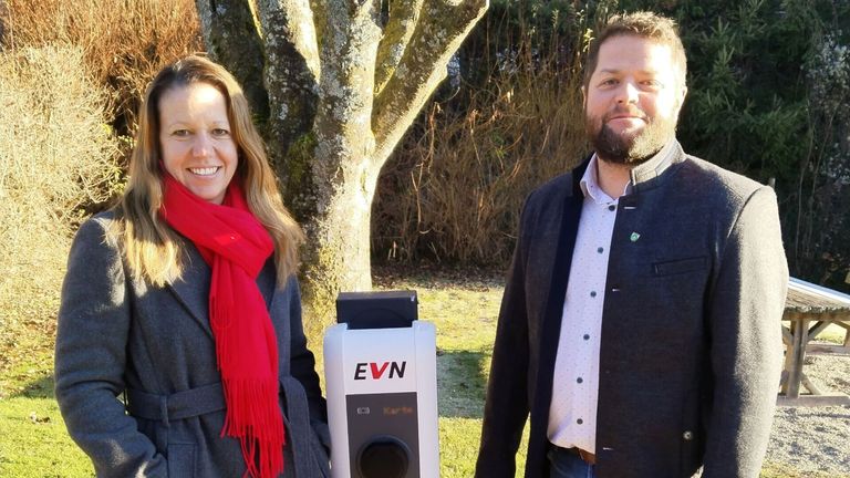 Ein Mann und eine Frau stehen neben einer EVN-Ladestation auf einer Rasenfläche. Die Frau trägt einen roten Schal und der Mann einen Bart.