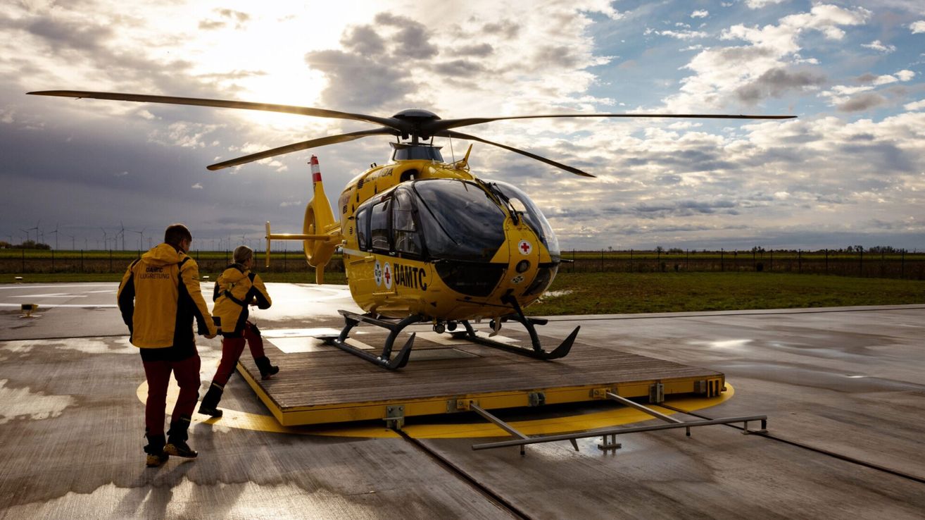 Ein gelber Hubschrauber mit der Aufschrift DAMTC steht auf dem Vorfeld, und eine Person in einer gelben Jacke geht darauf zu. Der Himmel ist teilweise bewölkt.