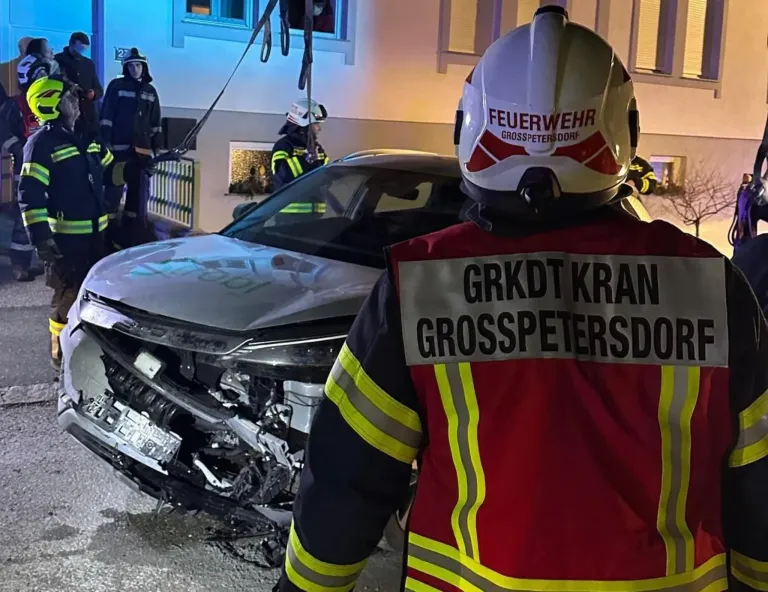 Feuerwehrleute stehen neben einem beschädigten Auto bei Nacht, einer trägt einen Helm mit dem Wort 'Feuerwehr'. Ein anderer steht bei einem Gebäude mit der Nummer 27.