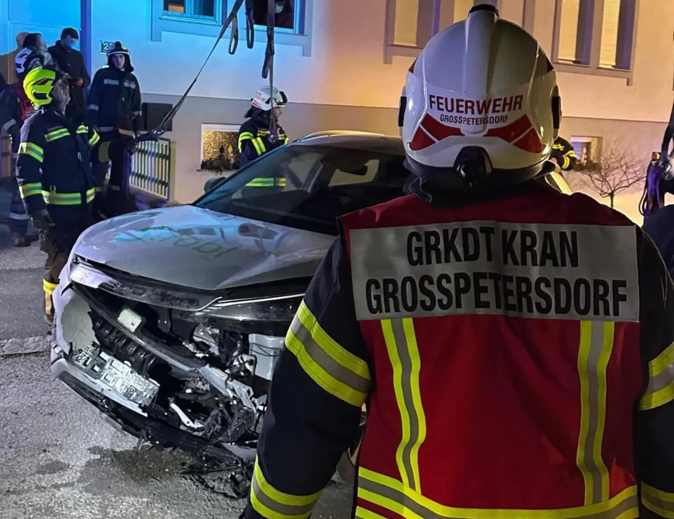 Feuerwehrleute stehen neben einem beschädigten Auto bei Nacht, einer trägt einen Helm mit dem Wort 'Feuerwehr'. Ein anderer steht bei einem Gebäude mit der Nummer 27.