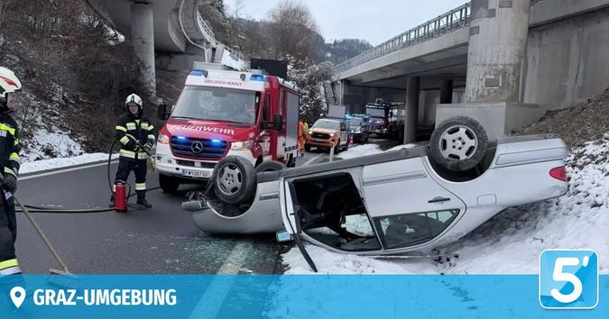 Ein Auto ist auf einer verschneiten Straße umgekippt, Feuerwehrleute sind am Unfallort. Ein Feuerlöscher steht in der Nähe. Ein roter und weißer Feuerwehrwagen ist hinter dem Auto geparkt.
