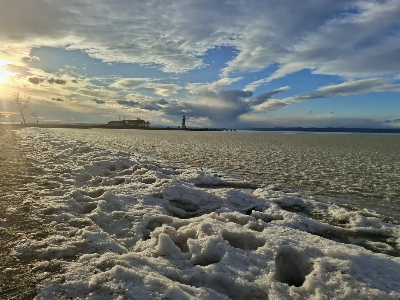 Ein gefrorener See mit schneebedecktem Eis und einem bewölkten Himmel. Ein Leuchtturm steht am entfernten Ufer.